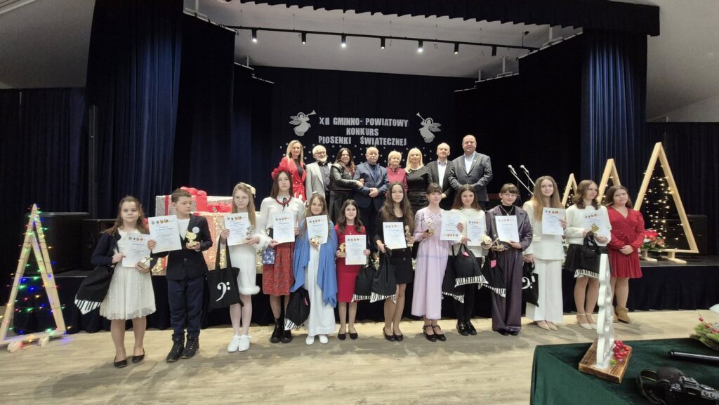 Uczestnicy konkursu z dyplomami i nagrodami za nimi na scenie jury, dyrektor szkoły i przedstawiciele urzędu gminy