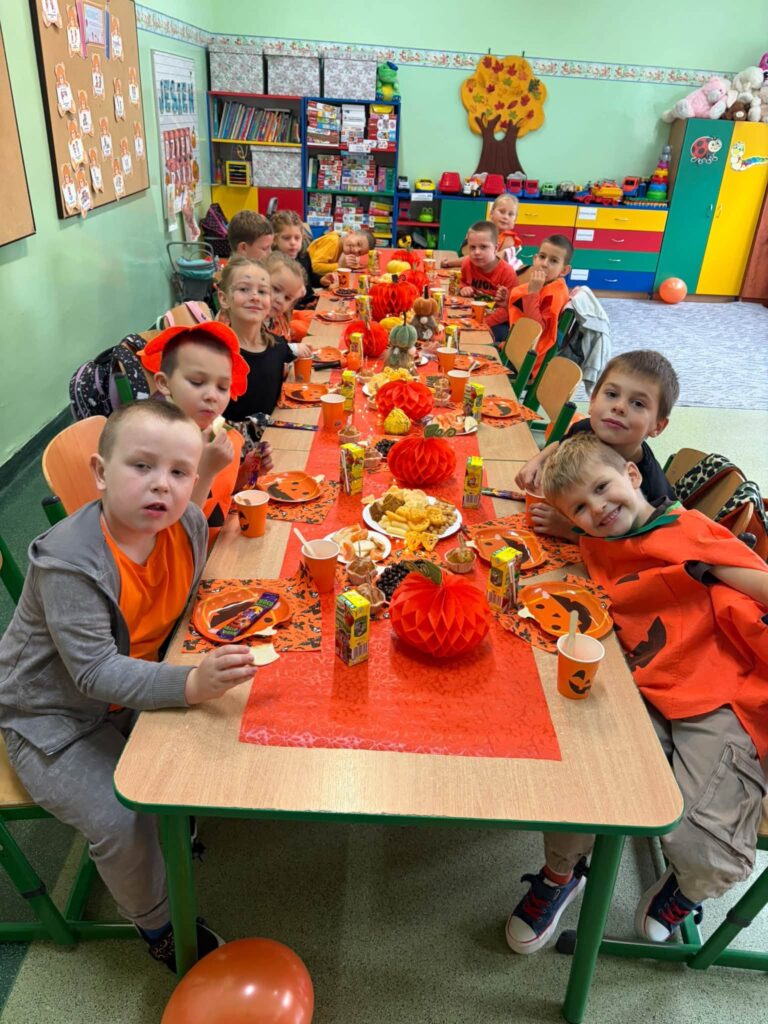 Przedszkolaki w pomarańczowych strojach siedzą przy stolikach z dyniowymi przekąskami